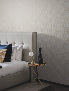 A modern bedroom featuring a gray upholstered bed adorned with blue and gold pillows, a round gold side table displaying a lamp, black bottle, and book, set against York Wallcoverings’ Serendipity Wallpaper in beige.