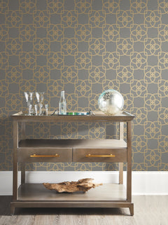 A modern bar cart with glassware, a bottle, a silver decorative orb, and a piece of driftwood sits against a gray wall adorned with York Wallcoverings’ Serendipity Wallpaper in a gold geometric pattern.