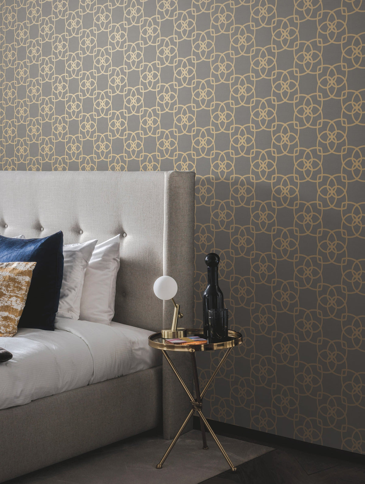 A modern bedroom featuring a gray tufted bed, gold and white pillows, and a small round gold table displaying a lamp, books, and decor, all set against York Wallcoverings’ Serendipity Wallpaper in a gray and gold geometric pattern.