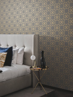 A modern bedroom featuring a gray tufted bed, gold and white pillows, and a small round gold table displaying a lamp, books, and decor, all set against York Wallcoverings’ Serendipity Wallpaper in a gray and gold geometric pattern.