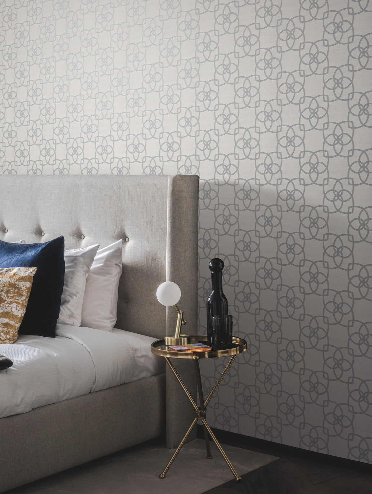 A modern bedroom features a tufted gray headboard, white and blue pillows, a gold round side table with a lamp and decorative bottle, set against York Wallcoverings’ Serendipity Wallpaper.