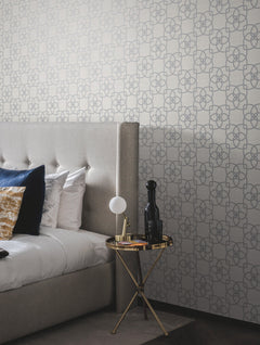 A modern bedroom features a tufted gray headboard, white and blue pillows, a gold round side table with a lamp and decorative bottle, set against York Wallcoverings’ Serendipity Wallpaper.