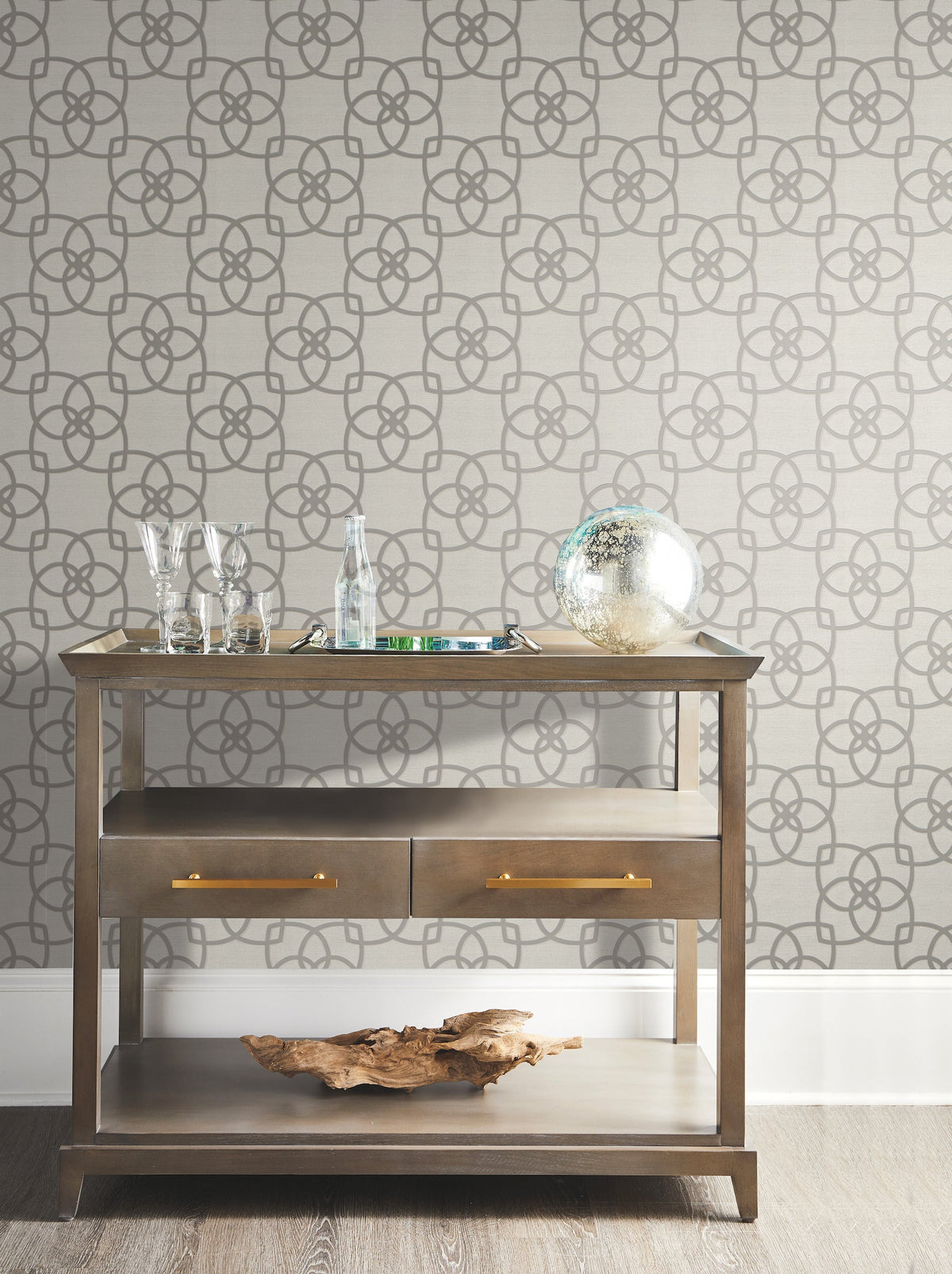 A wooden console table with glassware, a glass globe, and a driftwood piece sits against Serendipity Wallpaper by York Wallcoverings in a neutral-toned room.