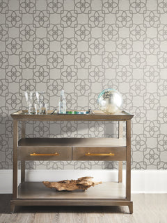 A wooden console table with glassware, a glass globe, and a driftwood piece sits against Serendipity Wallpaper by York Wallcoverings in a neutral-toned room.