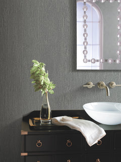 A modern bathroom featuring York Wallcoverings’ Natural Texture Wallpaper, paired with a black vanity cabinet with gold handles, a white bowl sink, wall-mounted faucet, a vase with green leaves, a folded towel, and a partial view of a mirror with a decorative border.