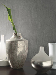A textured gray vase with a single green leaf stands beside a small, round, frosted silver vase on a dark surface against a wall covered in York Wallcoverings’ Natural Texture Wallpaper. White vases are visible in the background.