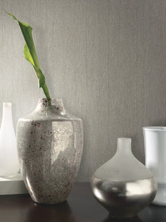 Two decorative vases are displayed on a dark surface. The larger vase features a speckled pattern and holds a single green leaf, while the smaller vase has a frosted and metallic finish. The background showcases York Wallcoverings’ Natural Texture Wallpaper, adding a neutral, textured look to the scene.