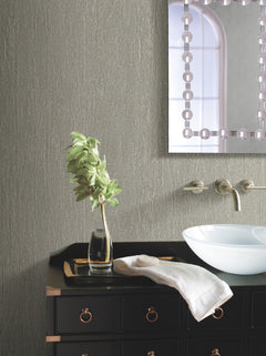 A modern bathroom vanity features York Wallcoverings’ Natural Texture Wallpaper as a backdrop, complemented by a black cabinet, gold drawer pulls, a glass vase with green leaves, a white towel, a vessel sink, wall-mounted faucets, and a mirror reflecting a window.