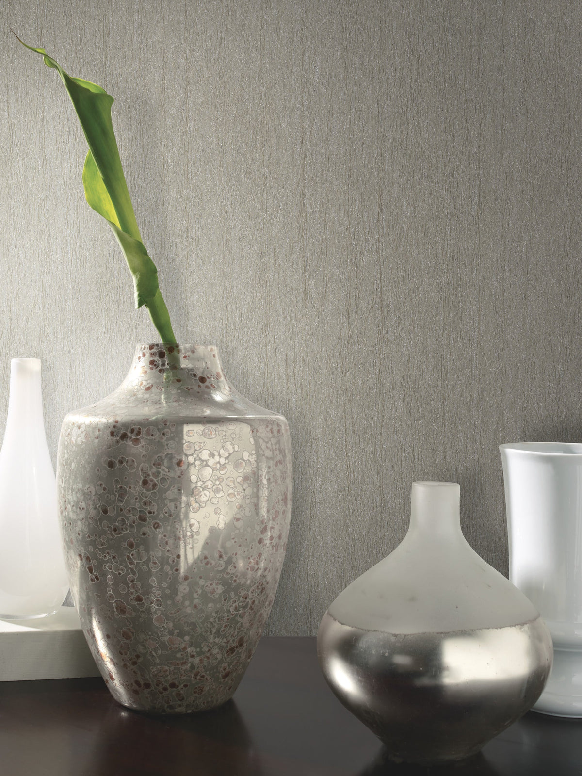 Three decorative vases sit on a dark surface against a wall adorned with Natural Texture Wallpaper by York Wallcoverings. The center vase is speckled with a single green leaf, the right vase is round and metallic, and the left vase is tall and white.