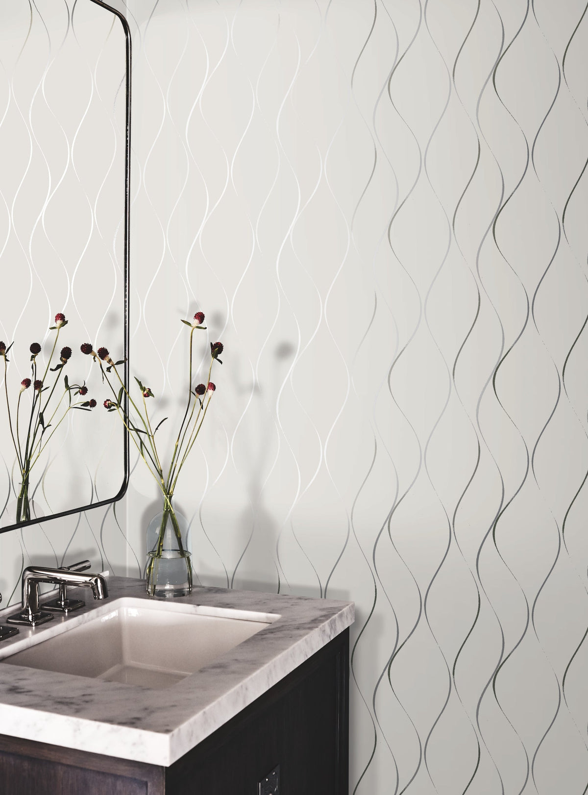 A bathroom vanity with a white sink, marble countertop, and a dark wood cabinet. A mirror and a glass vase with flowers sit on the counter. The wall features York Wallcoverings’ Wavy Stripe Wallpaper, adding a textured pattern to the space.