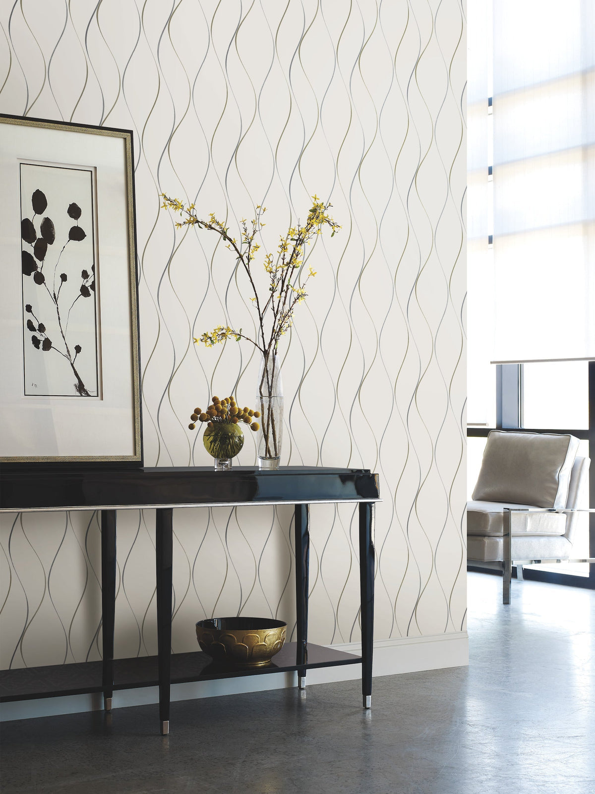 Modern interior with a black console table, a vase of yellow flowers, framed wall art, and a bowl. The look is completed by York Wallcoverings’ Wavy Stripe Wallpaper and a light-filled chair in the background.