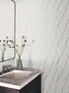 A bathroom sink with a marble countertop, a mirror, and a vase holding tall, thin flowers sits in front of a white wall featuring York Wallcoverings’ Wavy Stripe Wallpaper.