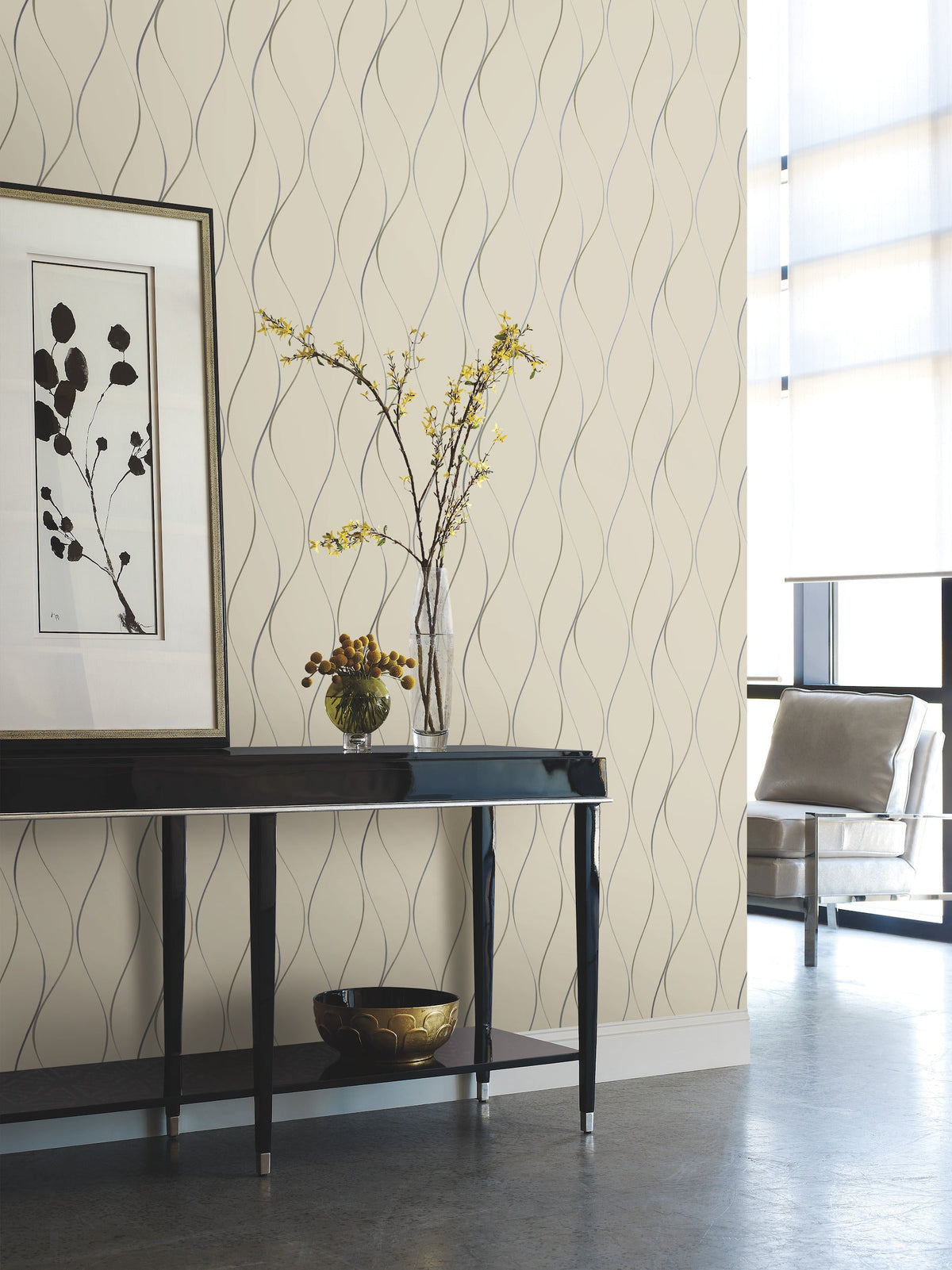 Modern living space featuring a black table, framed abstract art, a vase with yellow flowers, a decorative bowl, and a chair near large sunlit windows—all set against Wavy Stripe Wallpaper by York Wallcoverings.