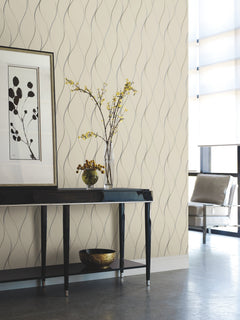Modern living space featuring a black table, framed abstract art, a vase with yellow flowers, a decorative bowl, and a chair near large sunlit windows—all set against Wavy Stripe Wallpaper by York Wallcoverings.