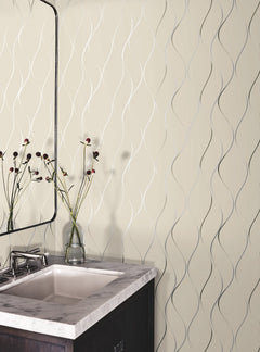 A bathroom featuring a marble countertop sink, a rectangular mirror, and a glass vase with flowers is accented by York Wallcoverings’ Wavy Stripe Wallpaper, which showcases a cream background with a silver wavy line pattern.