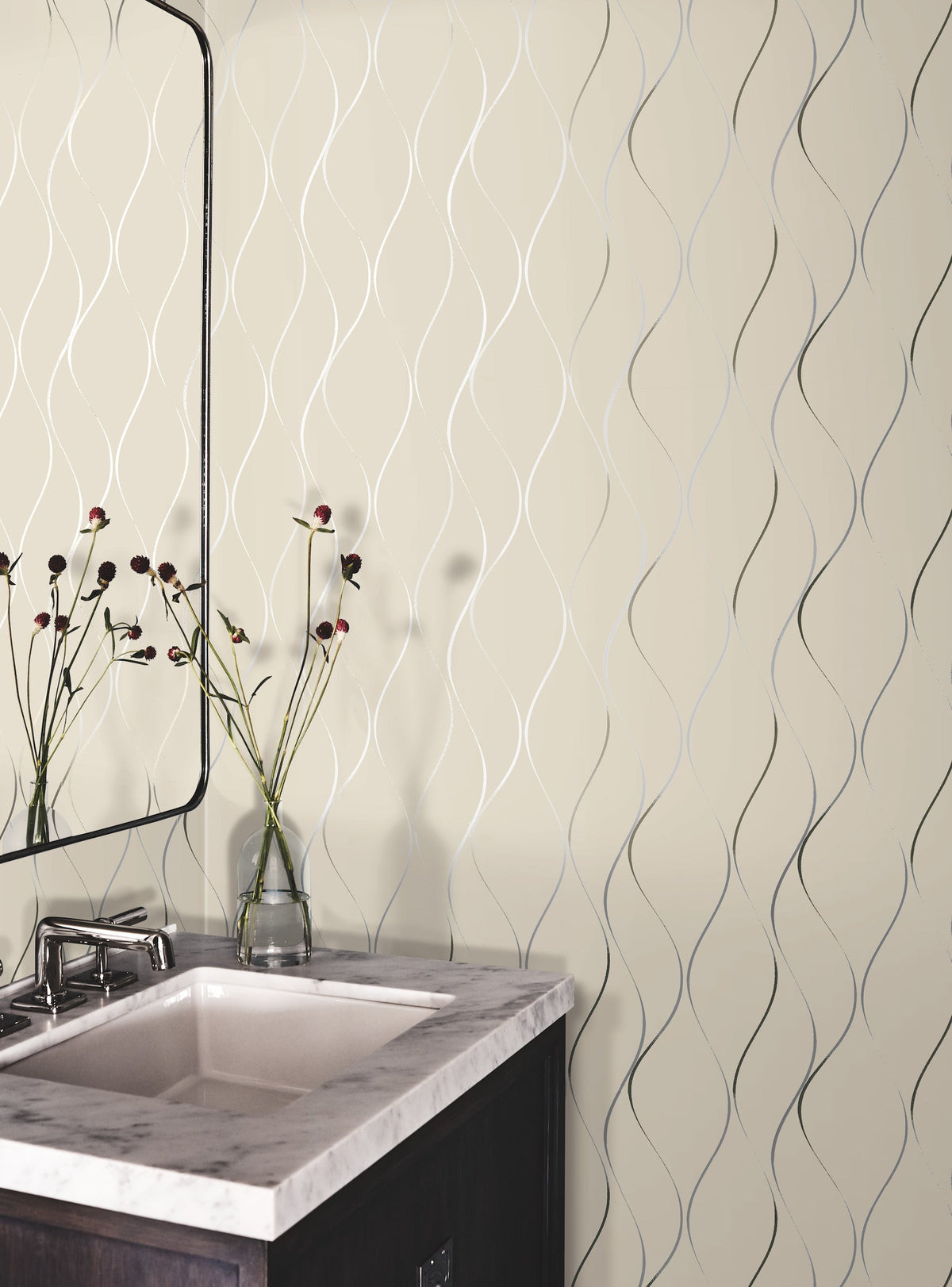A modern bathroom corner featuring a marble sink, a rectangular mirror, and a glass vase with long-stemmed flowers. The wall is adorned with Wavy Stripe Wallpaper by York Wallcoverings, showcasing a beige background accentuated by elegant wavy silver lines.