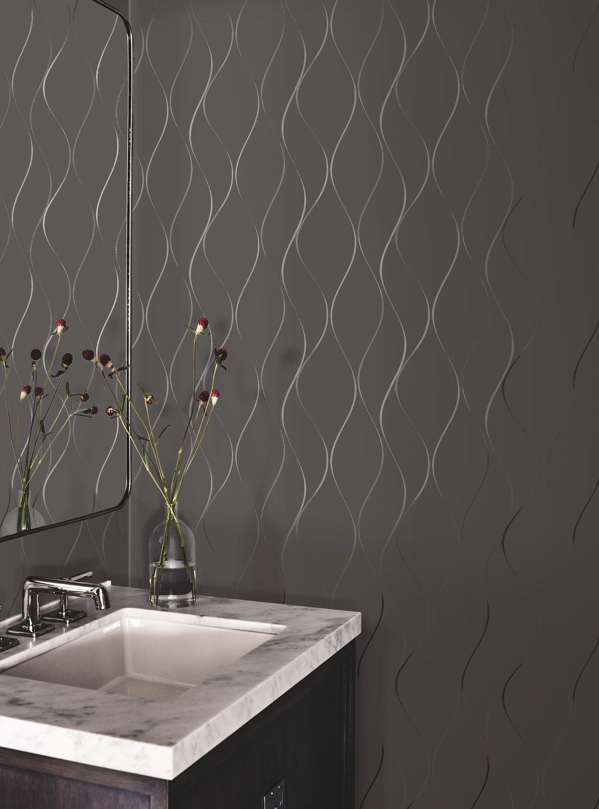 A modern bathroom featuring the Wavy Stripe Wallpaper by York Wallcoverings on a gray wall, complemented by a marble sink, a mirror, and a vase with long-stemmed flowers.