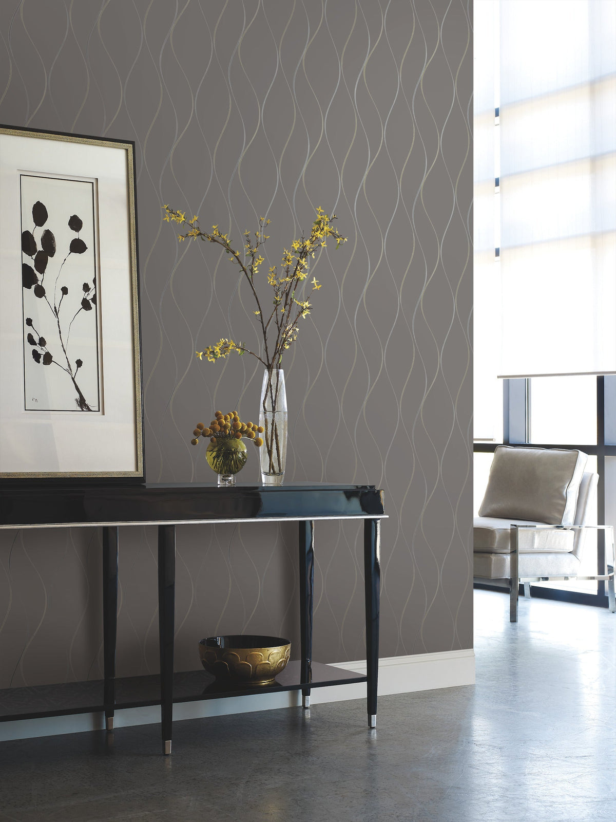 Modern living room featuring York Wallcoverings’ Wavy Stripe Wallpaper in gray, complemented by a black console table, framed botanical artwork, a glass vase with yellow flowers, a bowl underneath, and a metal chair positioned near large windows that fill the space with natural light.