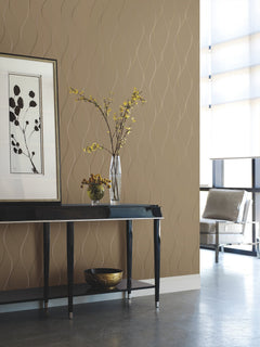A modern room featuring York Wallcoverings’ Wavy Stripe Wallpaper in beige, a black console table topped with a glass vase of yellow flowers, framed botanical art above, a bowl on the lower shelf, and a gray chair positioned near large windows.